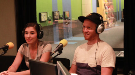 Two adults wearing headphones speaking into microphones in a radio studio