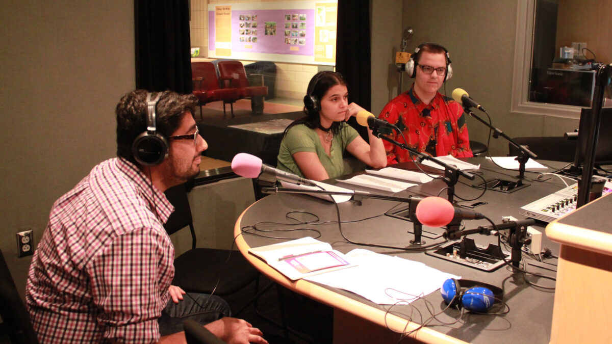 Three young adults wearing headphones and sitting behind microphones in a radio studio