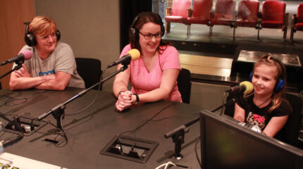 Two adults and a youth wearing headphones and speaking into microphones in a radio studio