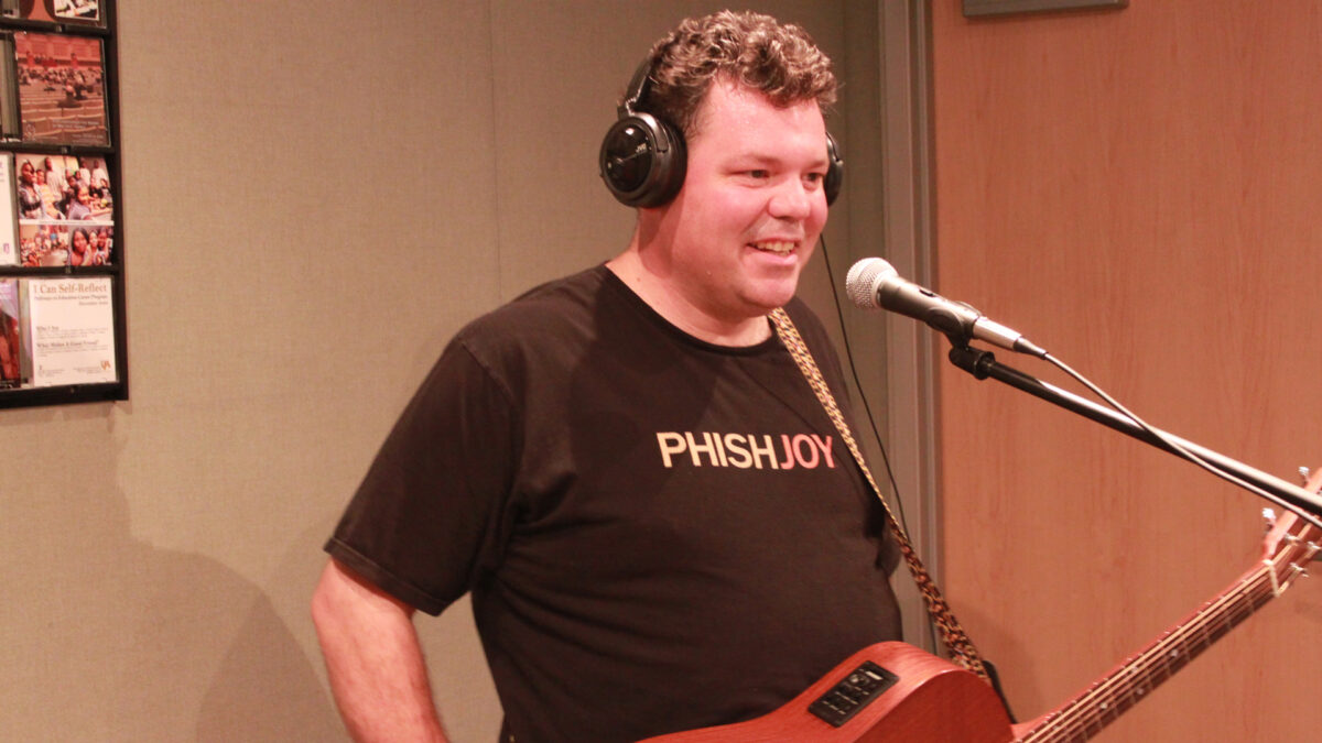 An adult wearing headphones smiling while holding an acoustic guitar and standing behind a microphone in a radio studio
