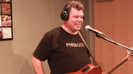 An adult wearing headphones smiling while holding an acoustic guitar and standing behind a microphone in a radio studio
