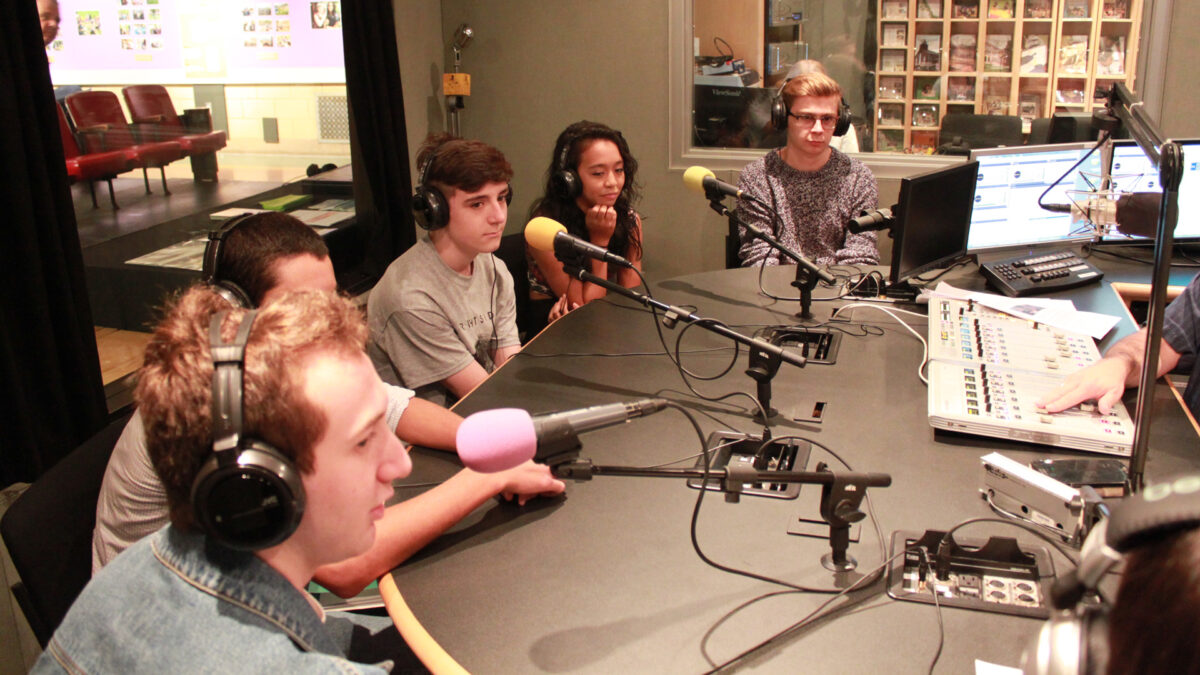 Five teens wearing headphones sitting behind microphones being interviewed in a radio studio