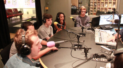 Five teens wearing headphones sitting behind microphones being interviewed in a radio studio