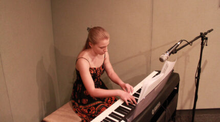 A teen playing a piano keyboard sitting behind sheet music and a microphone in a radio studio
