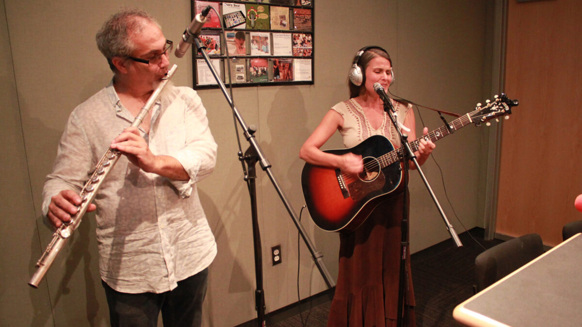 An adult playing a flute into a microphone and an adult wearing headphones playing an acoustic guitar and standing behind a microphone in a radio studio