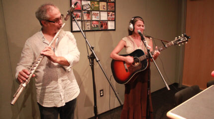 An adult playing a flute into a microphone and an adult wearing headphones playing an acoustic guitar and standing behind a microphone in a radio studio