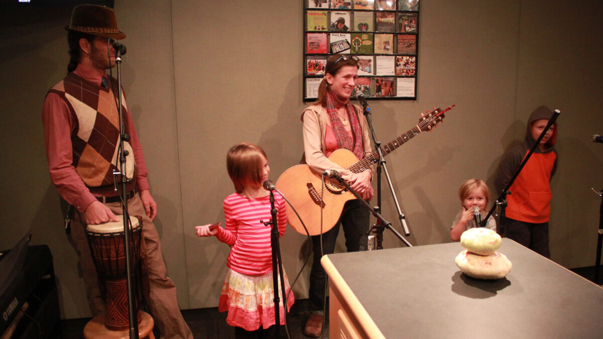 An adult standing behind a microphone with a hand on the head of a djembe, three youth standing behind microphones, and an adult holding an acoustic guitar standing behind a microphone in a radio studio