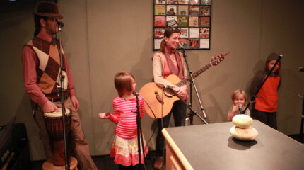 An adult standing behind a microphone with a hand on the head of a djembe, three youth standing behind microphones, and an adult holding an acoustic guitar standing behind a microphone in a radio studio