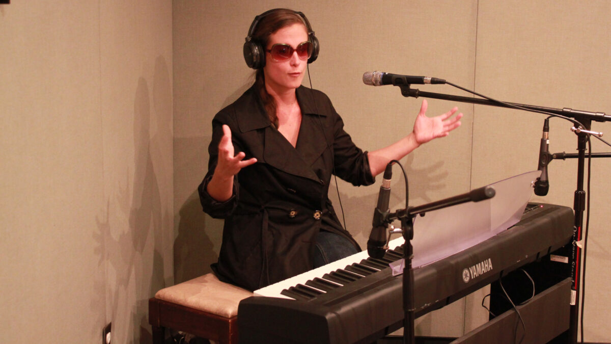 An adult wearing sunglasses and headphones sitting behind a piano keyboard and a microphone in a radio studio