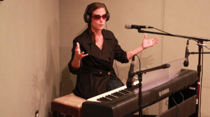 An adult wearing sunglasses and headphones sitting behind a piano keyboard and a microphone in a radio studio