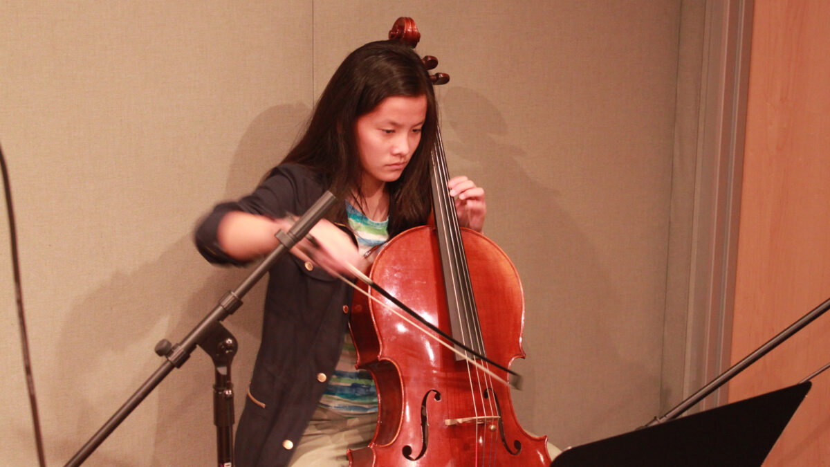 A youth playing cello looking at sheet music on a music stand in a radio studio