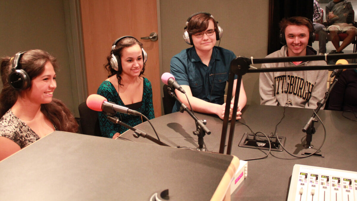 Four teens wearing headphones sitting behind microphones and smiling in a radio studio