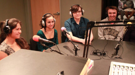 Four teens wearing headphones sitting behind microphones and smiling in a radio studio