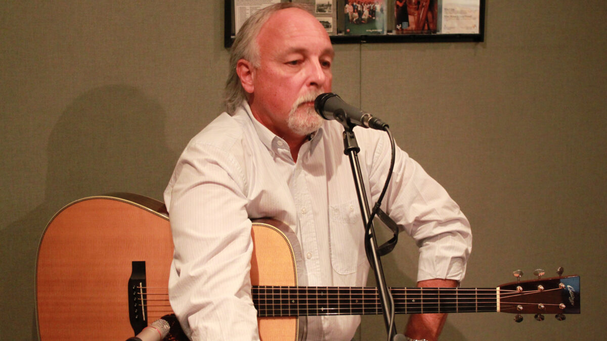 An adult holding an acoustic guitar standing behind a microphone in a radio studio