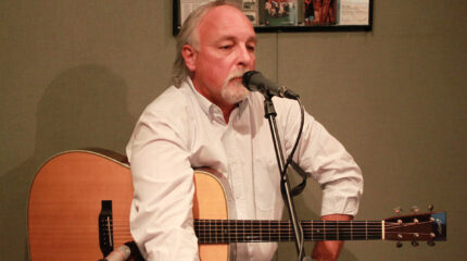 An adult holding an acoustic guitar standing behind a microphone in a radio studio