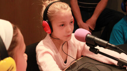 A youth wearing headphones looking at another youth wearing headphones speaking into a microphone in a radio studio