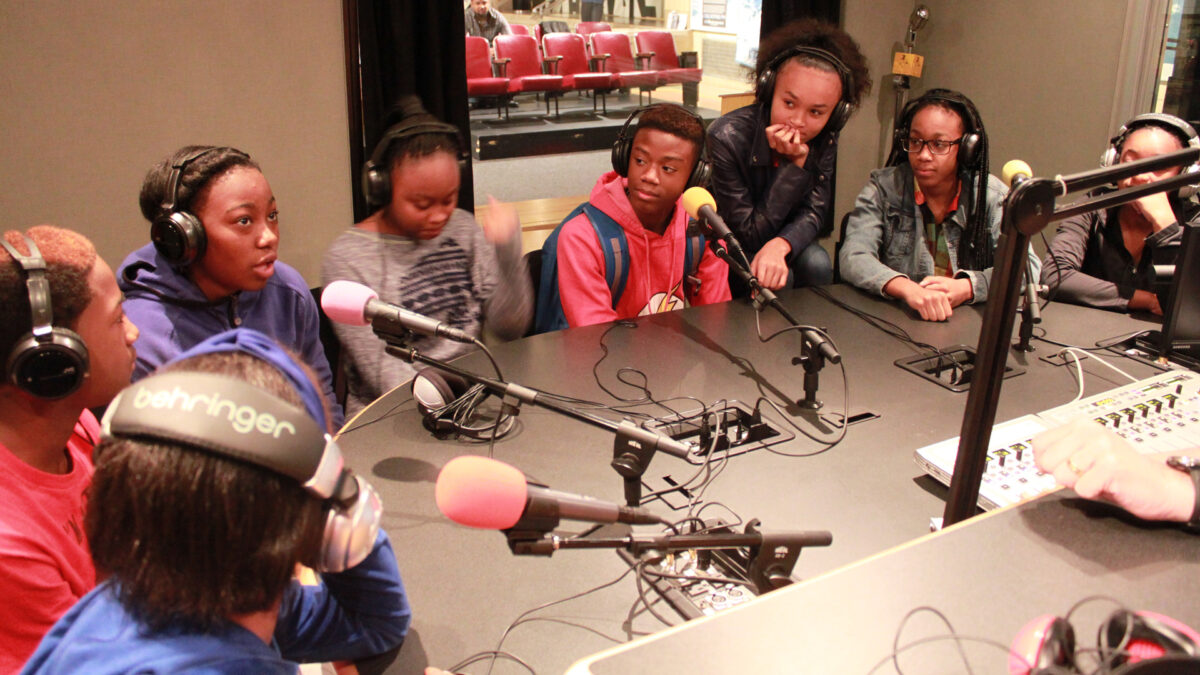 Eight youth wearing headphones speaking into microphones in a radio studio