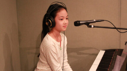 A youth wearing headphones sitting behind a piano keyboard and a microphone in a radio studio