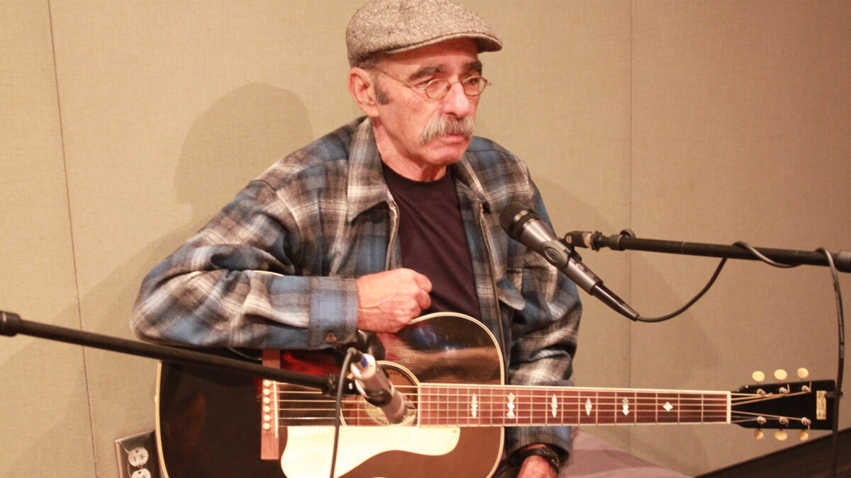 An adult holding an acoustic guitar sitting behind two microphones in a radio studio