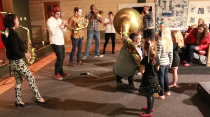 An adult playing alto saxophone, an adult playing trumpet, an adult playing French horn, two adults playing trombone, and an adult crouching in front of a small crowd of children while playing sousaphone
