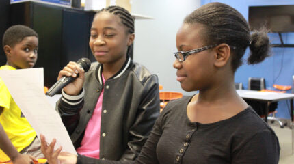 Two youth looking at a piece of paper, one holding a microphone, and another youth looking at them in a classroom