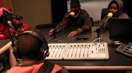 Four youth wearing headphones, three sitting behind microphones and one sitting behind a mixer, in a radio studio