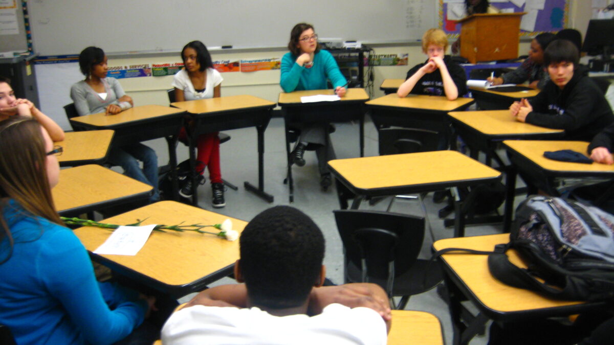 Eight youth sitting at desks in a circle in a classroom in front of a whiteboard