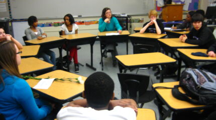 Eight youth sitting at desks in a circle in a classroom in front of a whiteboard