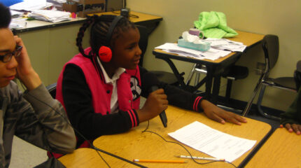 Two youth sitting at classroom desks, one wearing headphones and speaking into a microphone