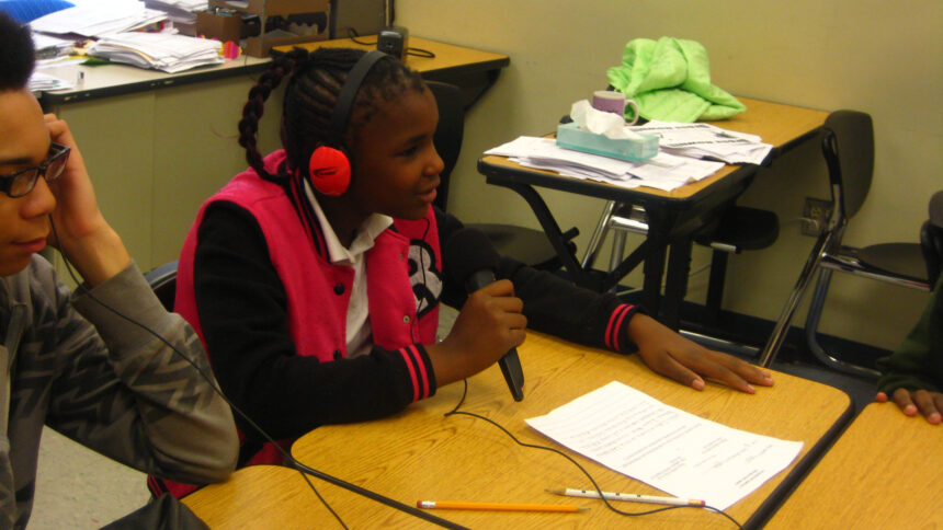 Two youth sitting at classroom desks, one wearing headphones and speaking into a microphone