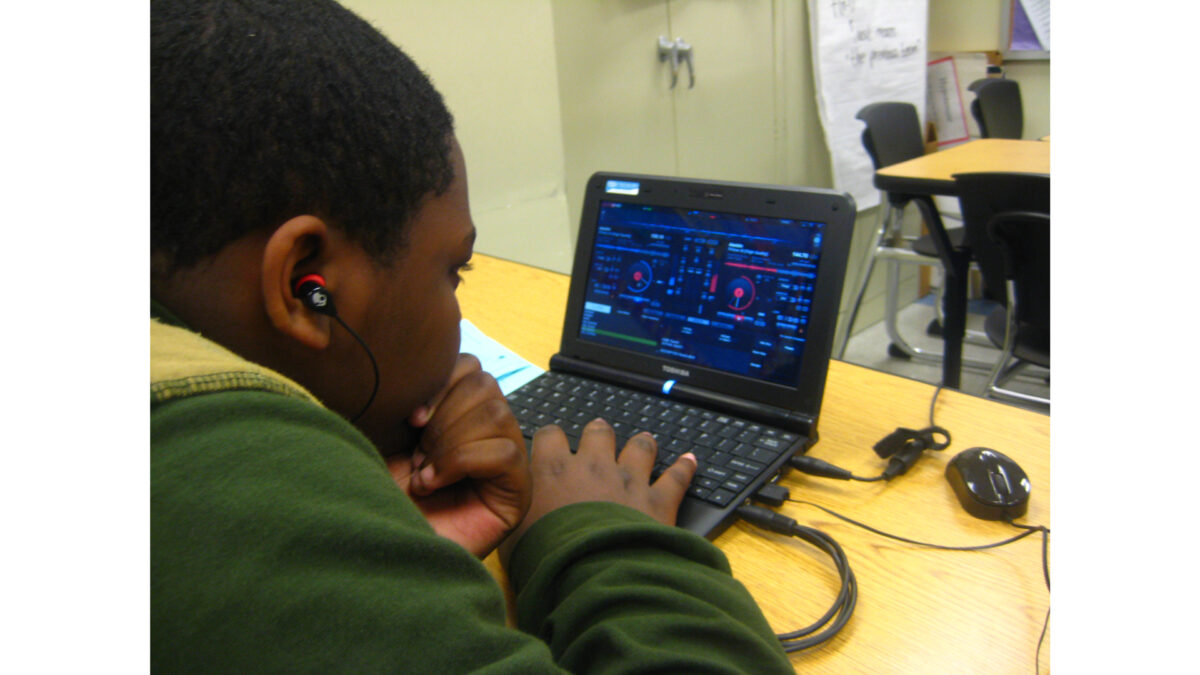 A youth wearing earbuds looking at audio software on a laptop computer on a classroom desk