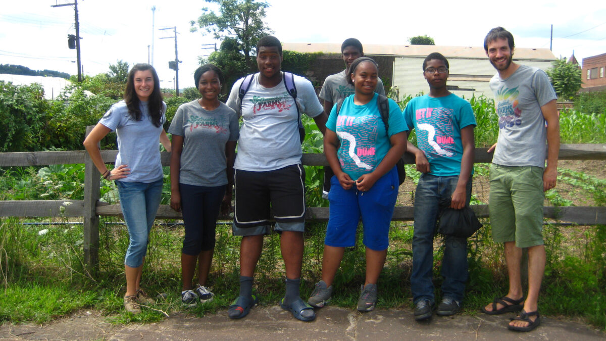 Six teens and one adult smiling and standing in front of a brown wooden fence around a small garden