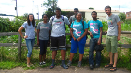 Six teens and one adult smiling and standing in front of a brown wooden fence around a small garden