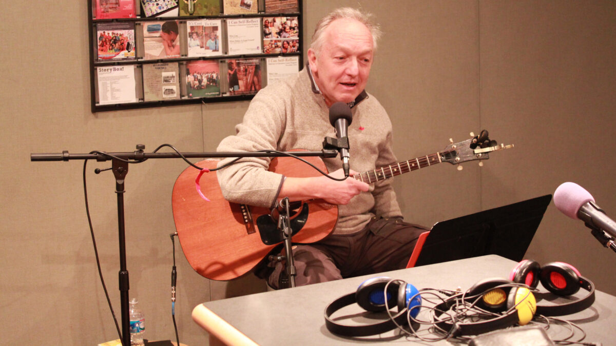 An adult holding an acoustic guitar and speaking into a microphone sitting behind a music stand in a radio studio