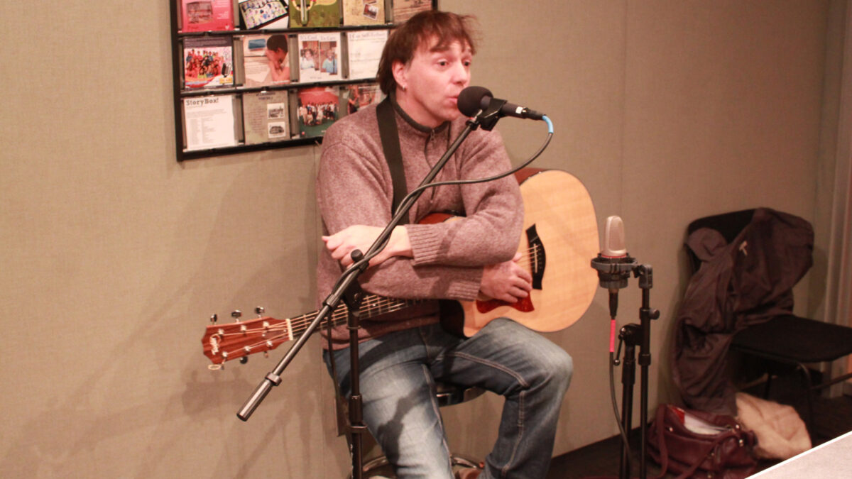 An adult holding an acoustic guitar and speaking into a microphone in a radio studio