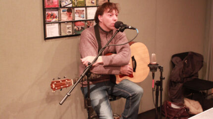An adult holding an acoustic guitar and speaking into a microphone in a radio studio