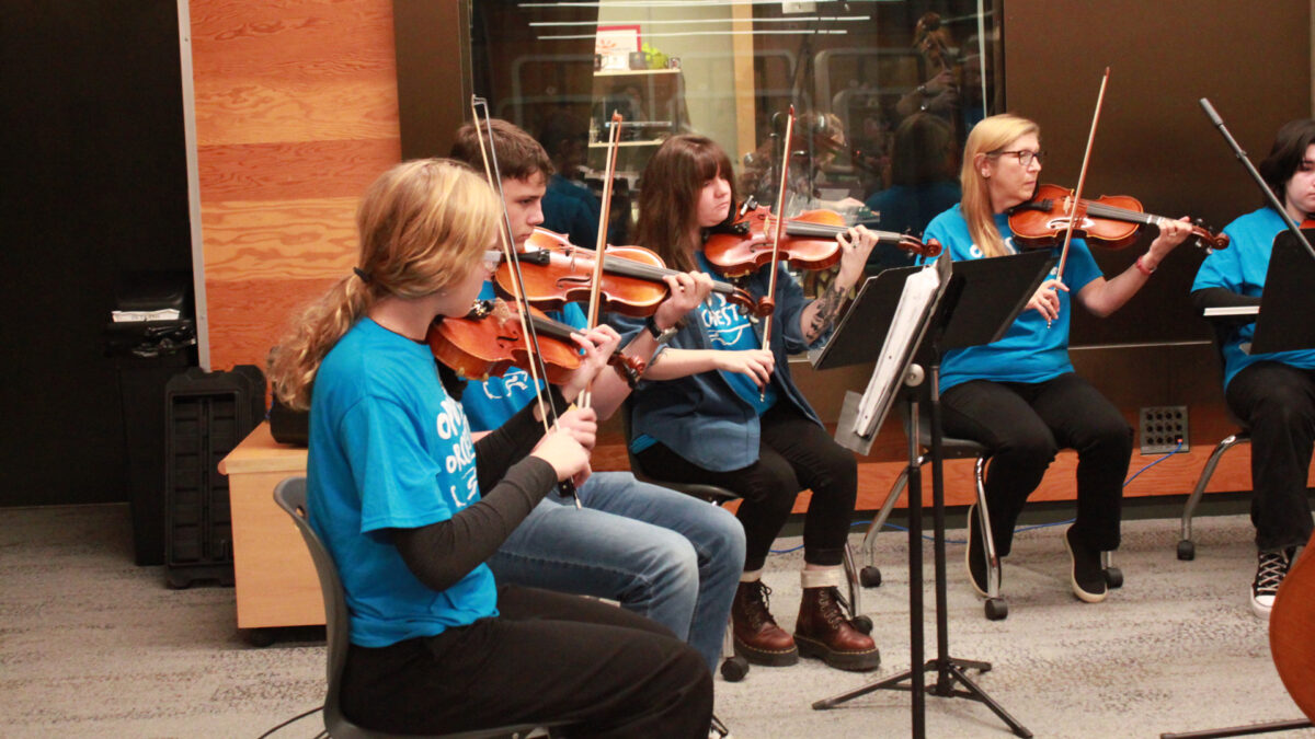 Four individuals playing violins in an orchestra sitting behind sheet music on music stands