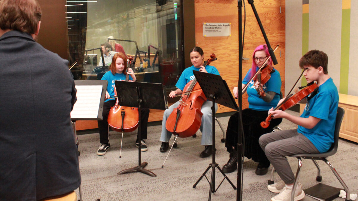 Two individuals playing violas and two individuals playing cellos in an orchestra sitting behind sheet music on music stands in a radio studio