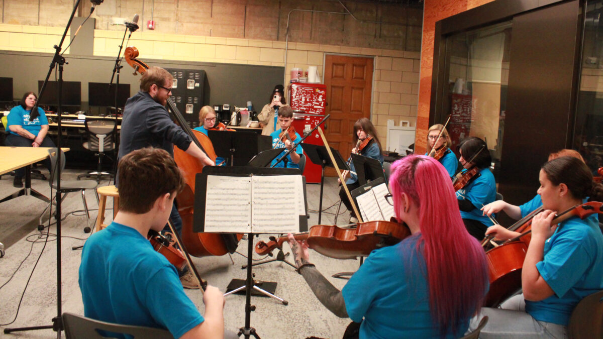 Five individuals playing violins, two individuals playing cellos, two individuals playing violas, and an individual playing upright bass in an orchestra sitting behind sheet music on music stands in a radio studio