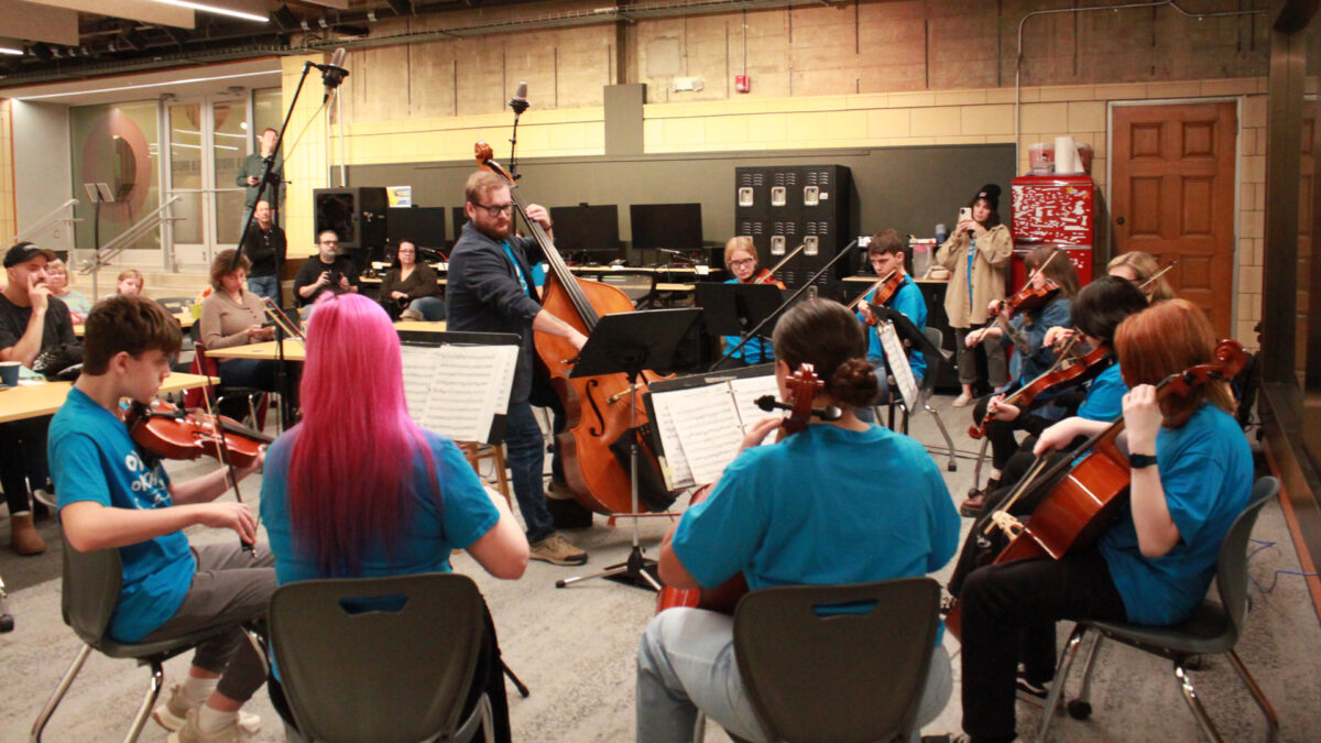 Five individuals playing violins, two individuals playing cellos, two individuals playing violas, and an individual playing an upright bass in an orchestra sitting behind sheet music on music stands as eight individuals listen in the background