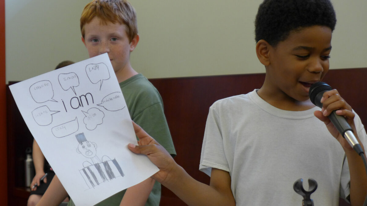 A youth speaking into a microphone while holding a piece of paper as another youth watches