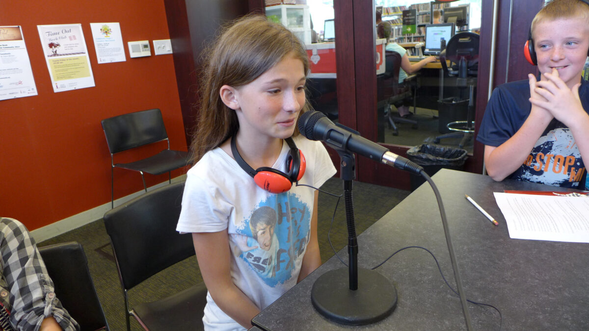 A youth wearing headphones around their neck speaking into a microphone and a youth wearing headphones listening