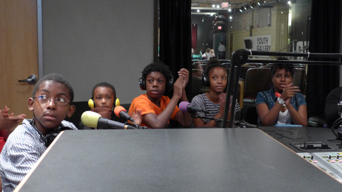 Five youth wearing headphones sitting behind microphones in a radio studio