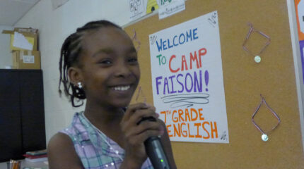 A youth smiling while holding a microphone and standing in front of a cork board in a classroom