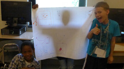 Two youth posing next to a youth holding a large piece of poster paper in front of them casting them in shadow in a classroom