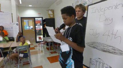 An adult looking at a youth holding a piece of paper and speaking into a microphone in front of a white board as four youth watch while sitting at classroom desks