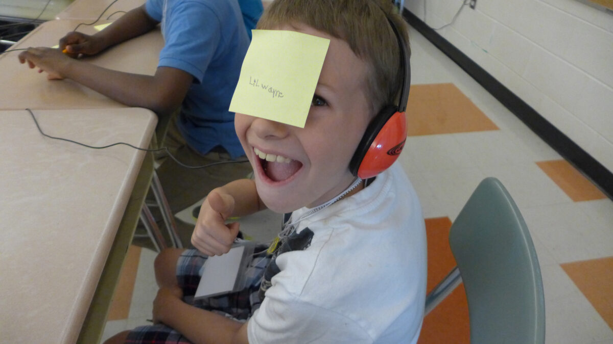 A youth with a sticky note on their face and wearing headphones smiling widely sitting at a classroom desk