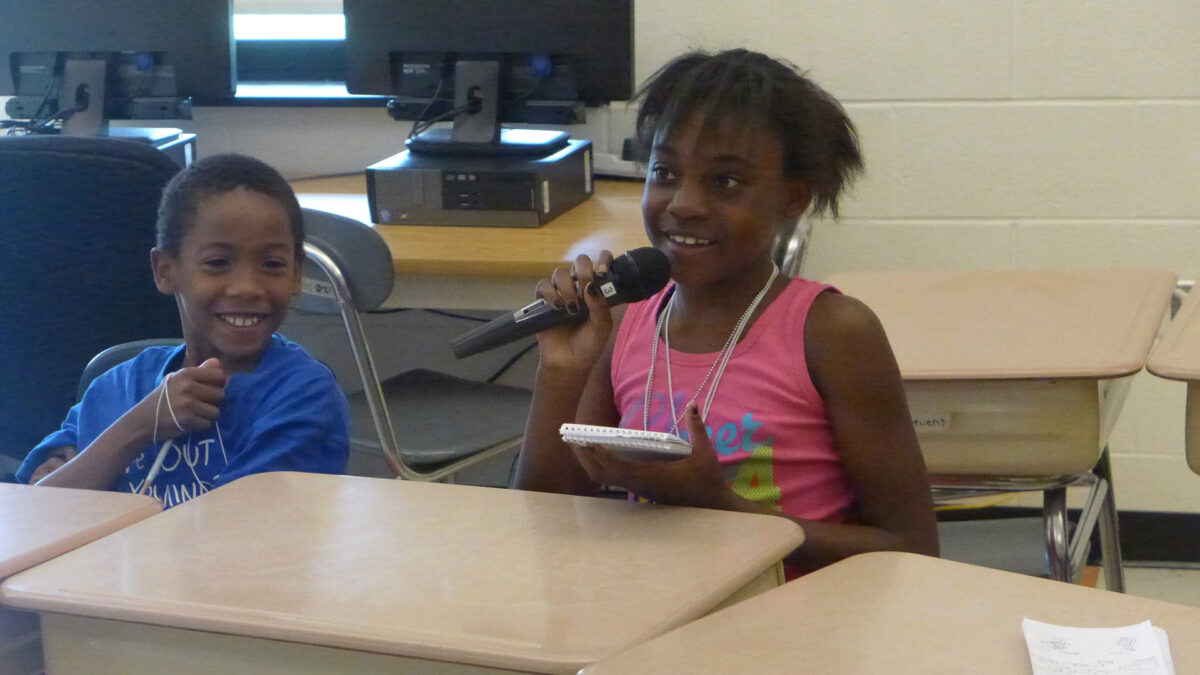 Two youth sitting at classroom desks, one holding a notepad and speaking into a microphone