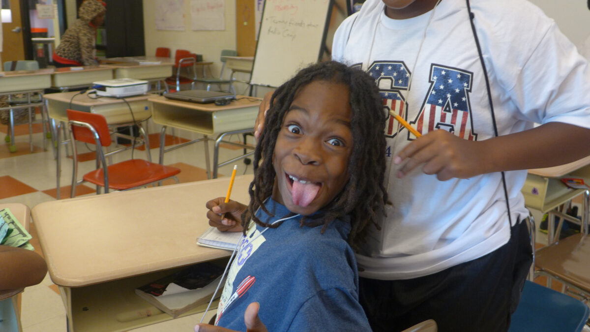 A youth making a silly face at the camera while sitting at a classroom desk