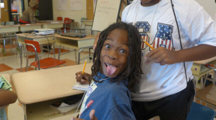 A youth making a silly face at the camera while sitting at a classroom desk